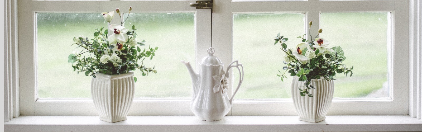 A decorative tea kettle flanked by two matching potted plants resting upon a sunny window sill